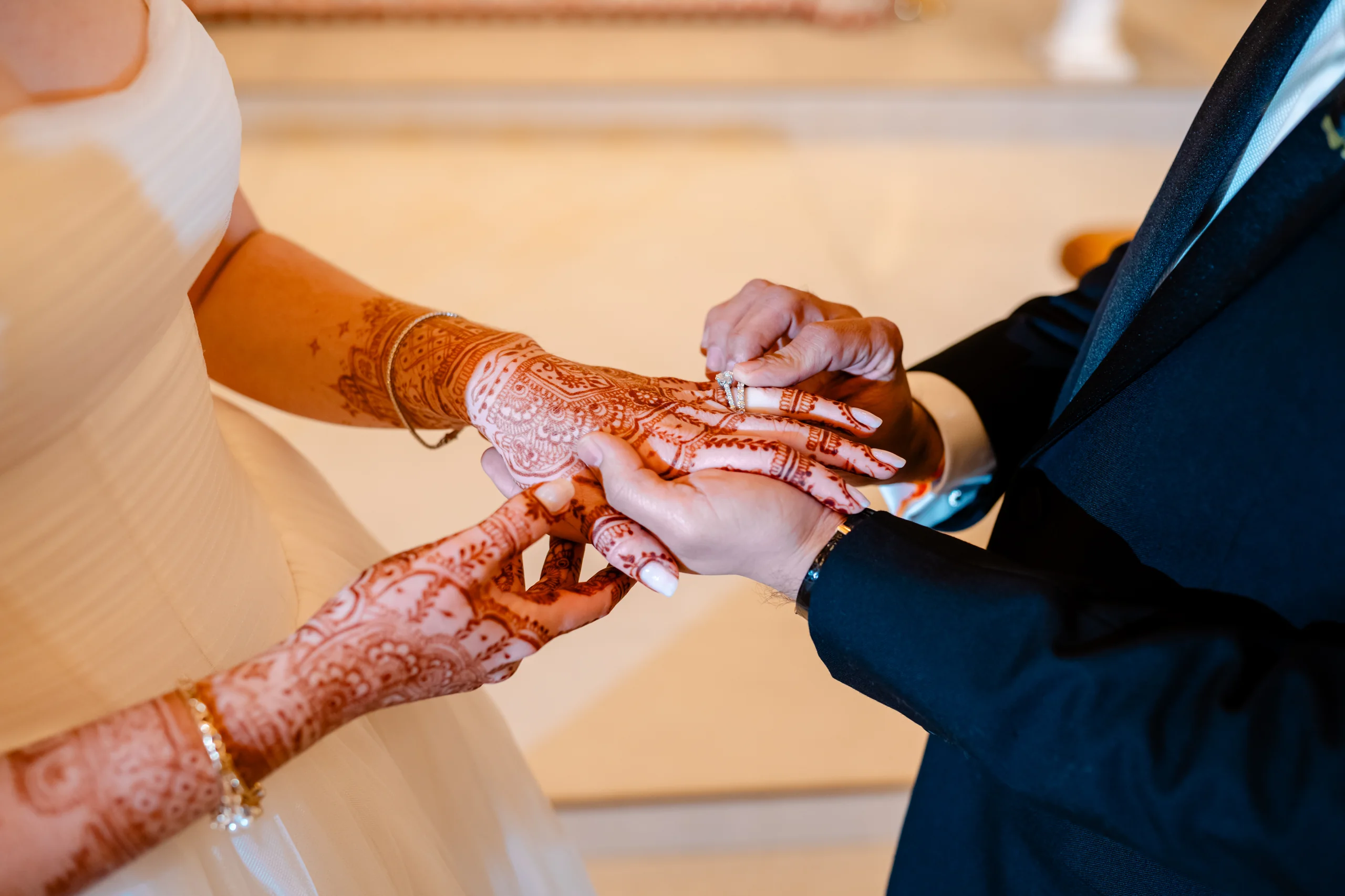 A groom in a tuxedo placing a wedding ring on his bride's hand adorned with intricate henna and mehndi art