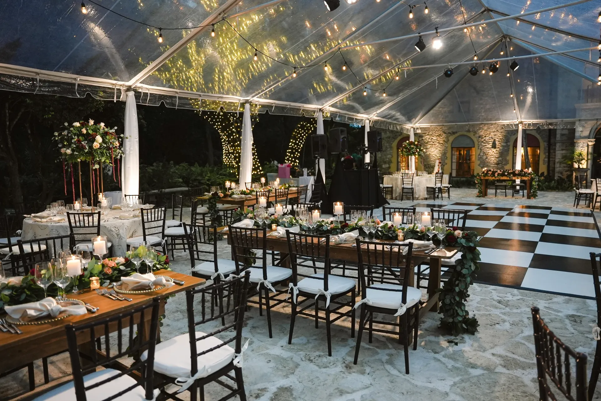 Clear-top tented wedding reception illuminated with bistro lights, checkered dance floor and elegant floral arrangements
